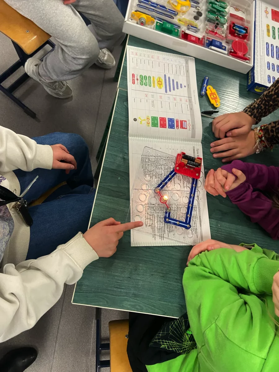 Kolejna grupa uczniów pochyla się nad książką ze schematem jakiegoś obwodu elektrycznego. Na książce leży płytka montażowa a na niej zamocowany jest koszyk z ogniwami, do której podłączona jest świecąca się żarówka.