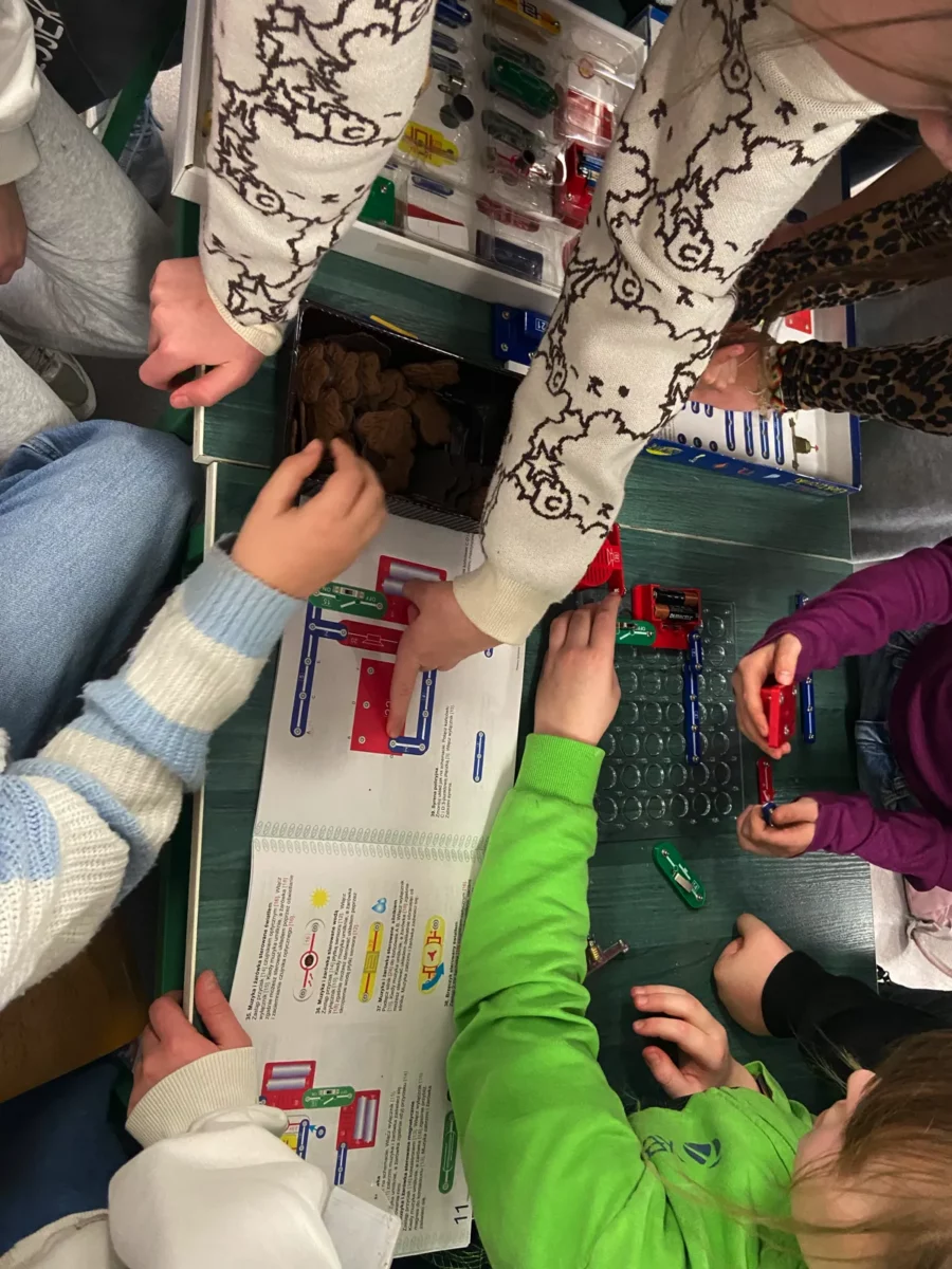 Uczniowie zgromadzeni wokół płytki montażowej z elementami elektronicznymi. Jeden z nich wskazuje na jakiś element na schemacie elektrycznym wydrukowanym w książce.