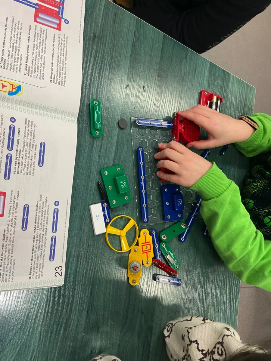 Uczeń składa kolejne urządzenie. Na płytce umieścił kilka łączników, teraz mocuje głośniczek. Obok na stole leżą inne elementy, m.in. wiatraczek, silniczek, przełącznik, element światłoczuły itd.
